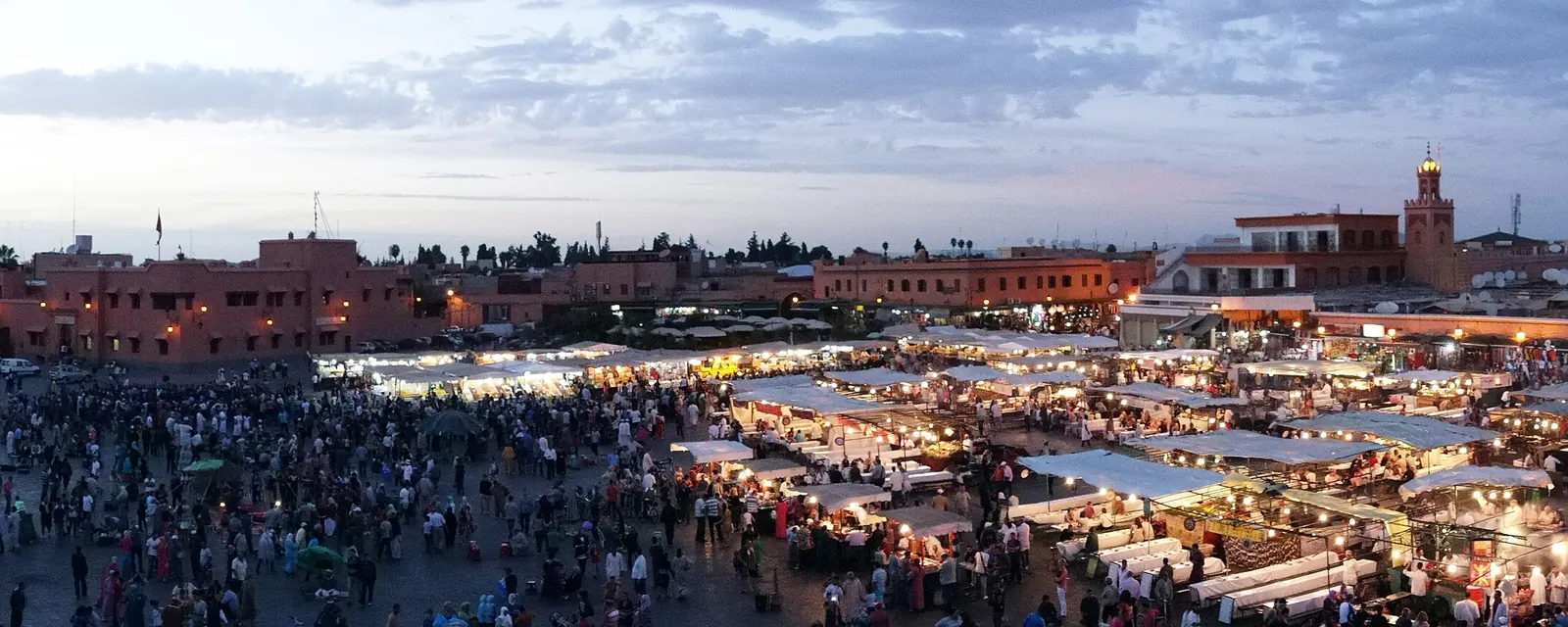A wide panorama of the Jemaa el-Fnaa square in Marrakesh in the evening, with the crowd, stalls and the Koutoubia minaret in the distance