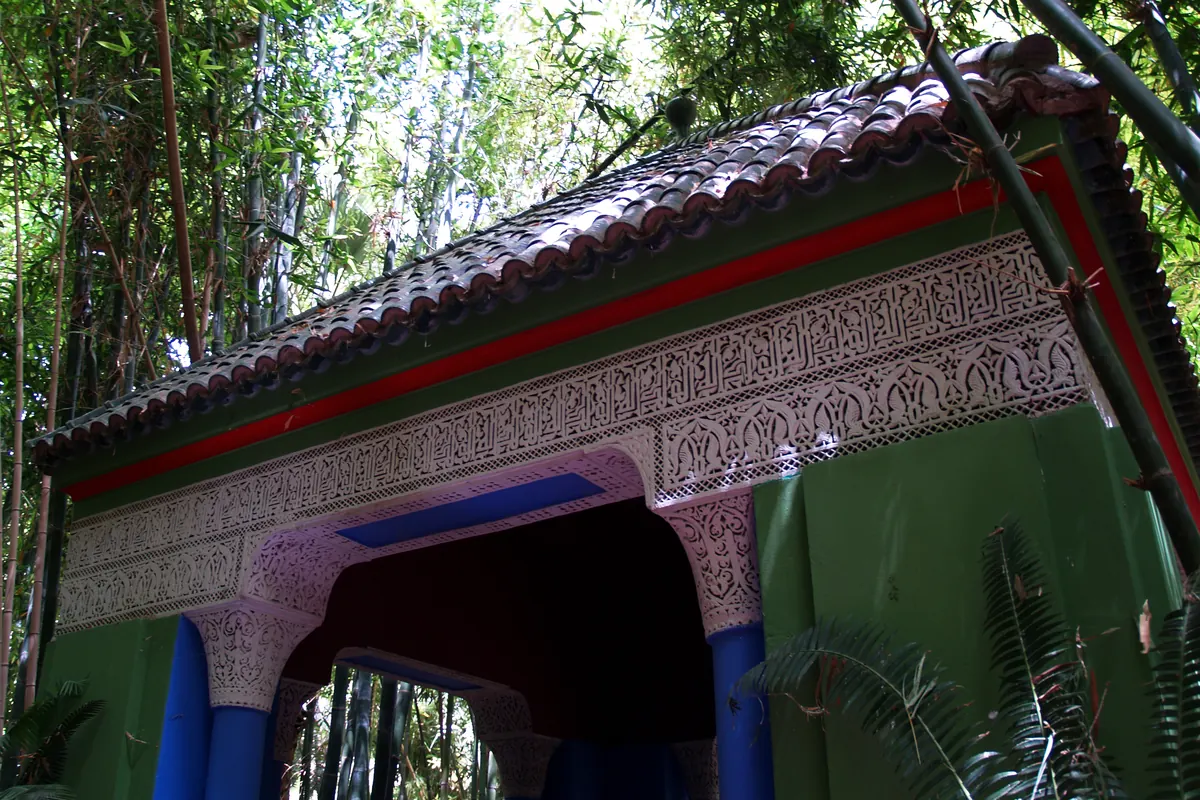 A green painted wooden pavilion with a carved plaster frieze and a tiled roof, with bamboo behind it in the Majorelle Garden