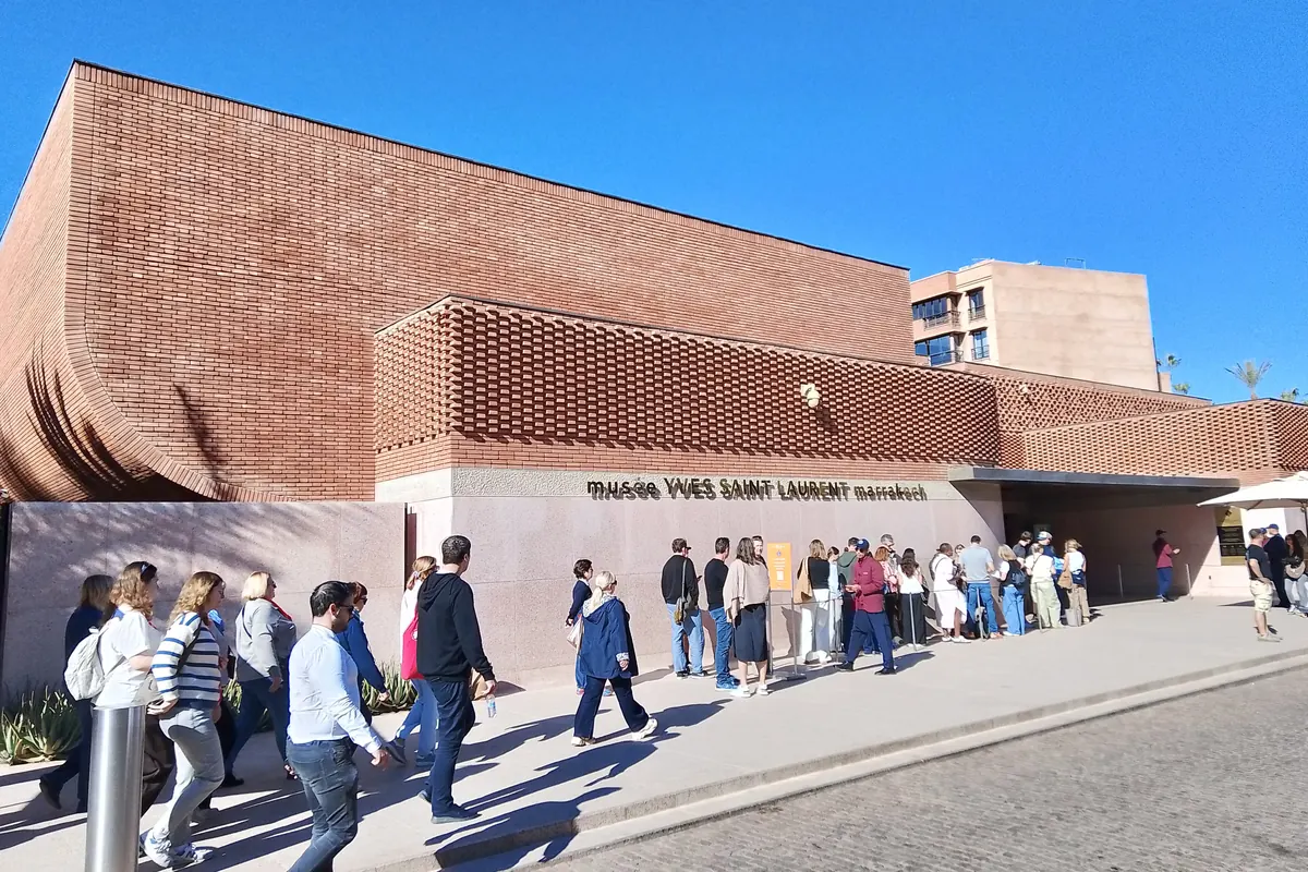 The brick facade of the Musée Yves Saint Laurent Marrakech under a blue sky, with a line of visitors along the pavement at its entrance